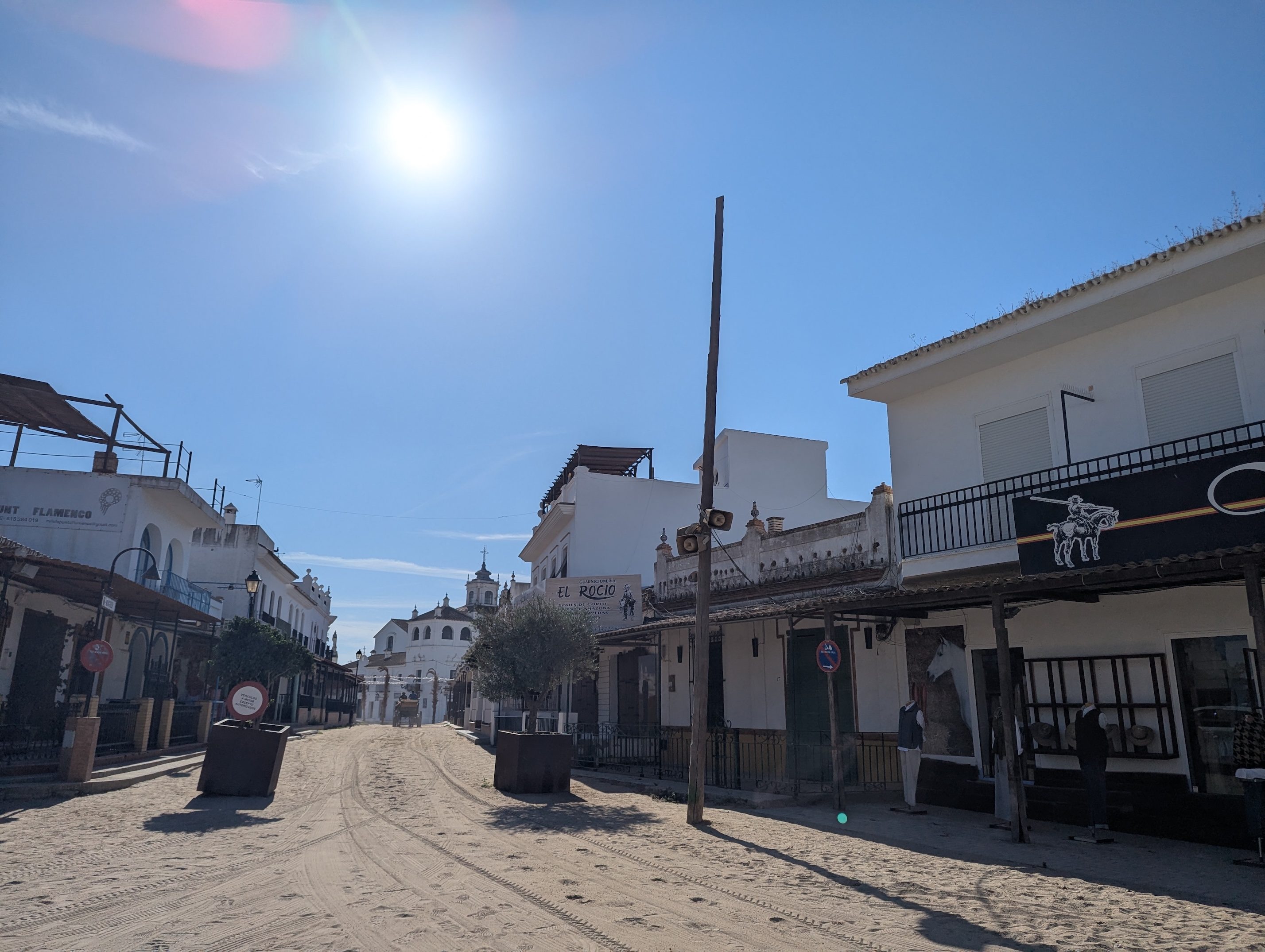 A wide sandy street in El Rocío with tire tracks, low white buildings with wooden balconies, and a church tower in the distance under a bright sun