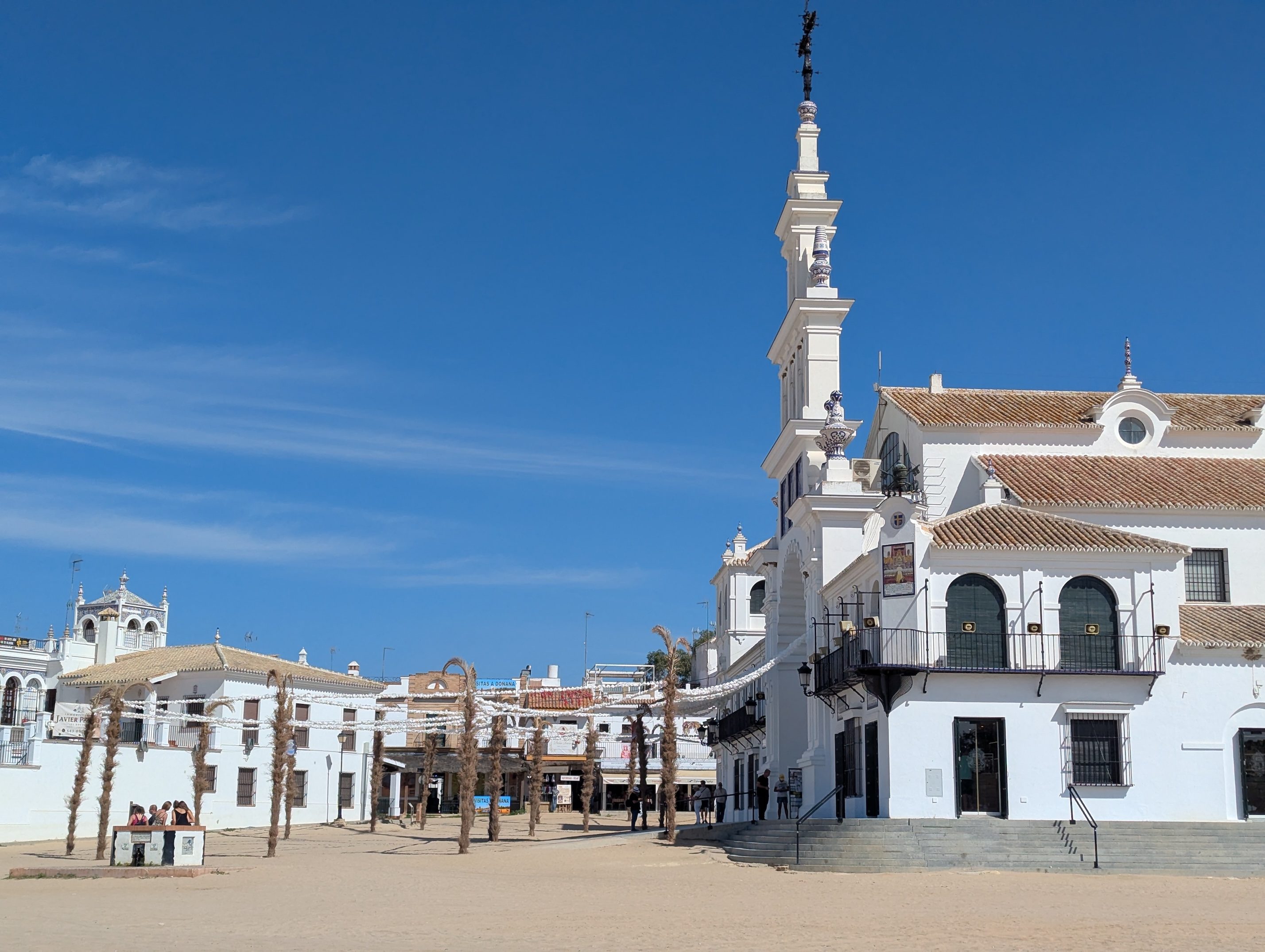 The white church and bell tower of El Rocío beside the wide sandy main square, with palm-trunk decorations and a blue sky