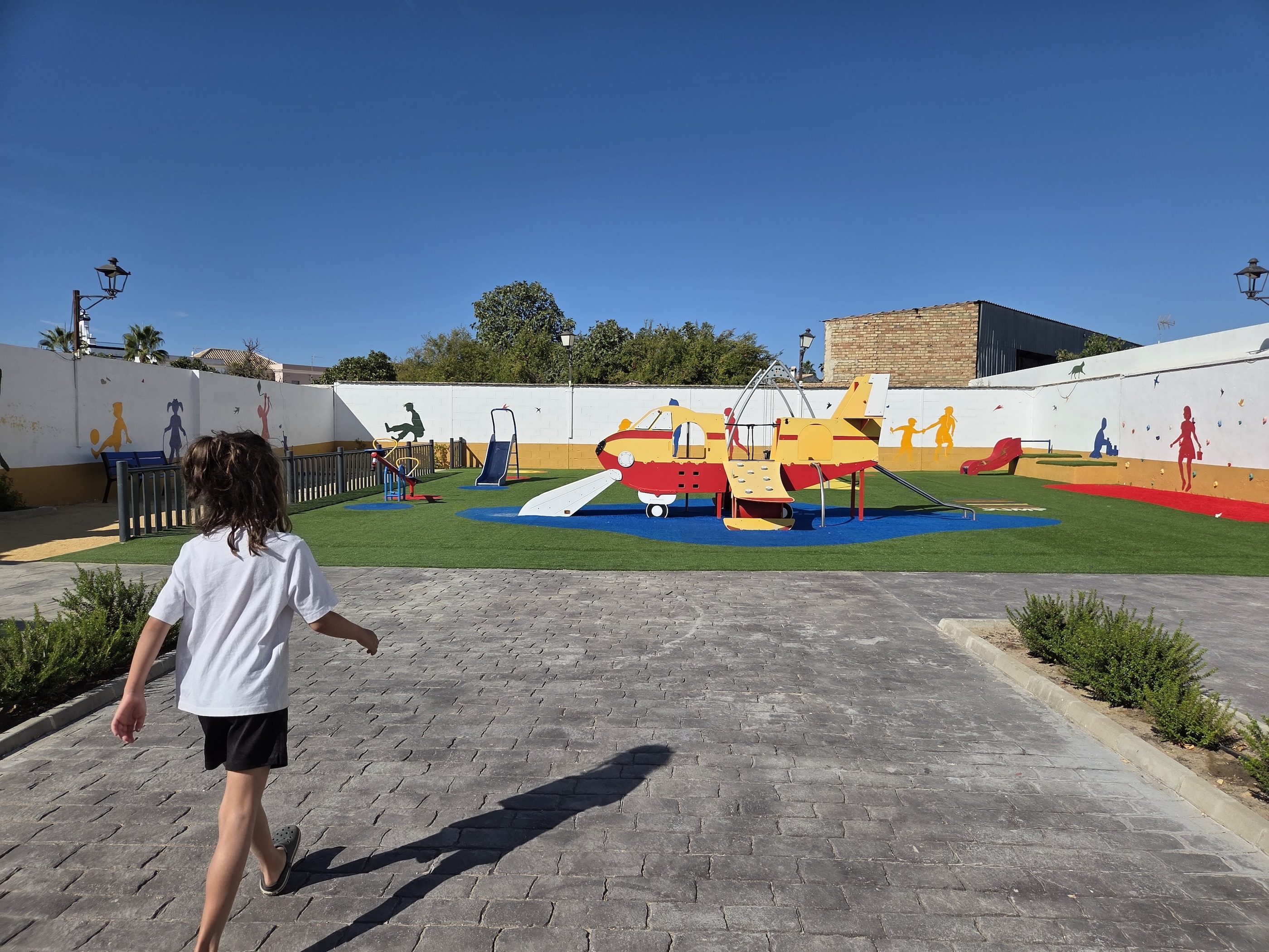 Remi walking toward a colorful airplane-shaped playground with a slide on bright artificial turf, a white wall painted with figures behind it, in Hinojos