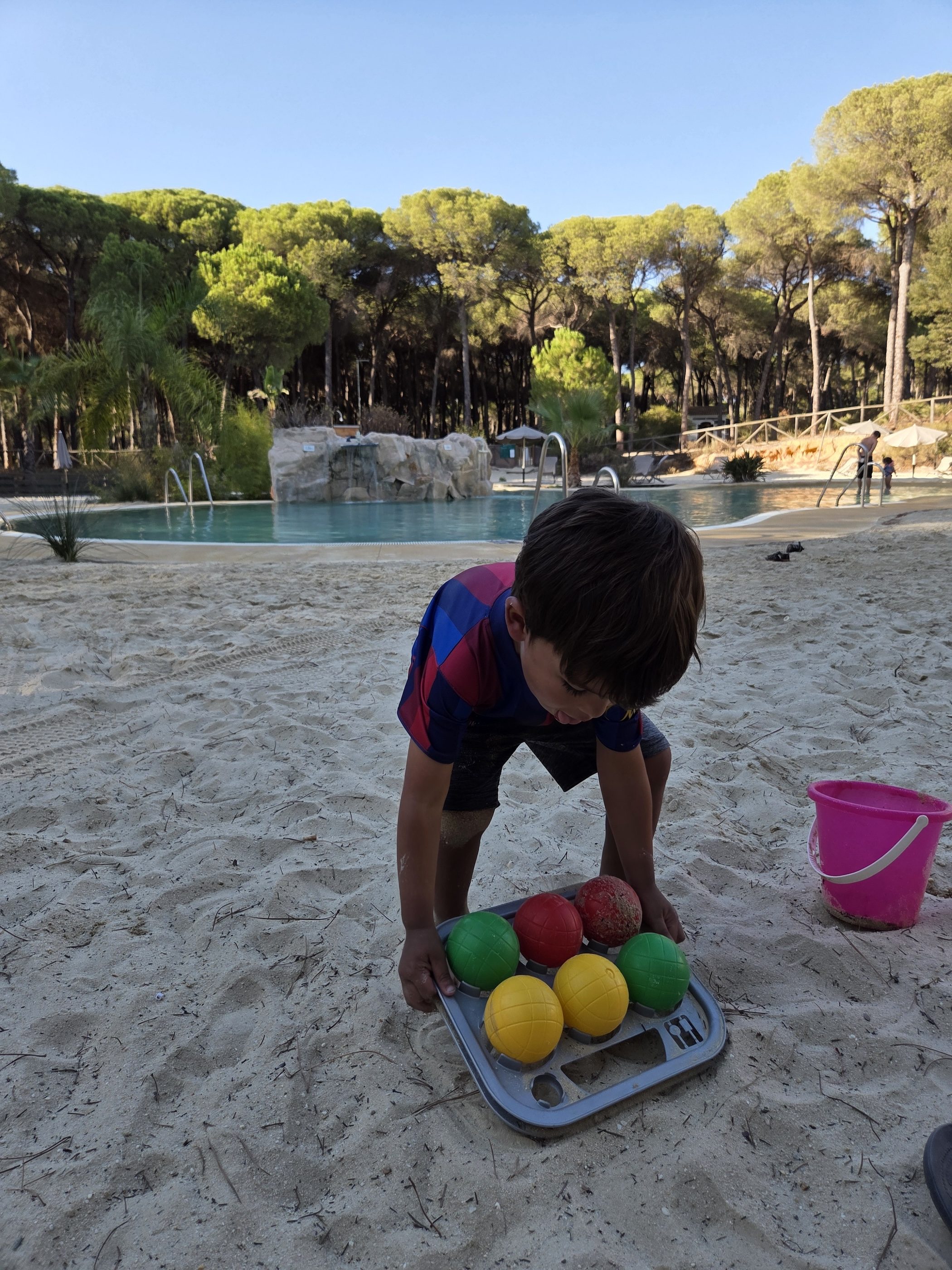 Arlo in his Barça jersey picking up a boules set of red, green and yellow balls from the sand, a pink bucket beside him and the pool behind