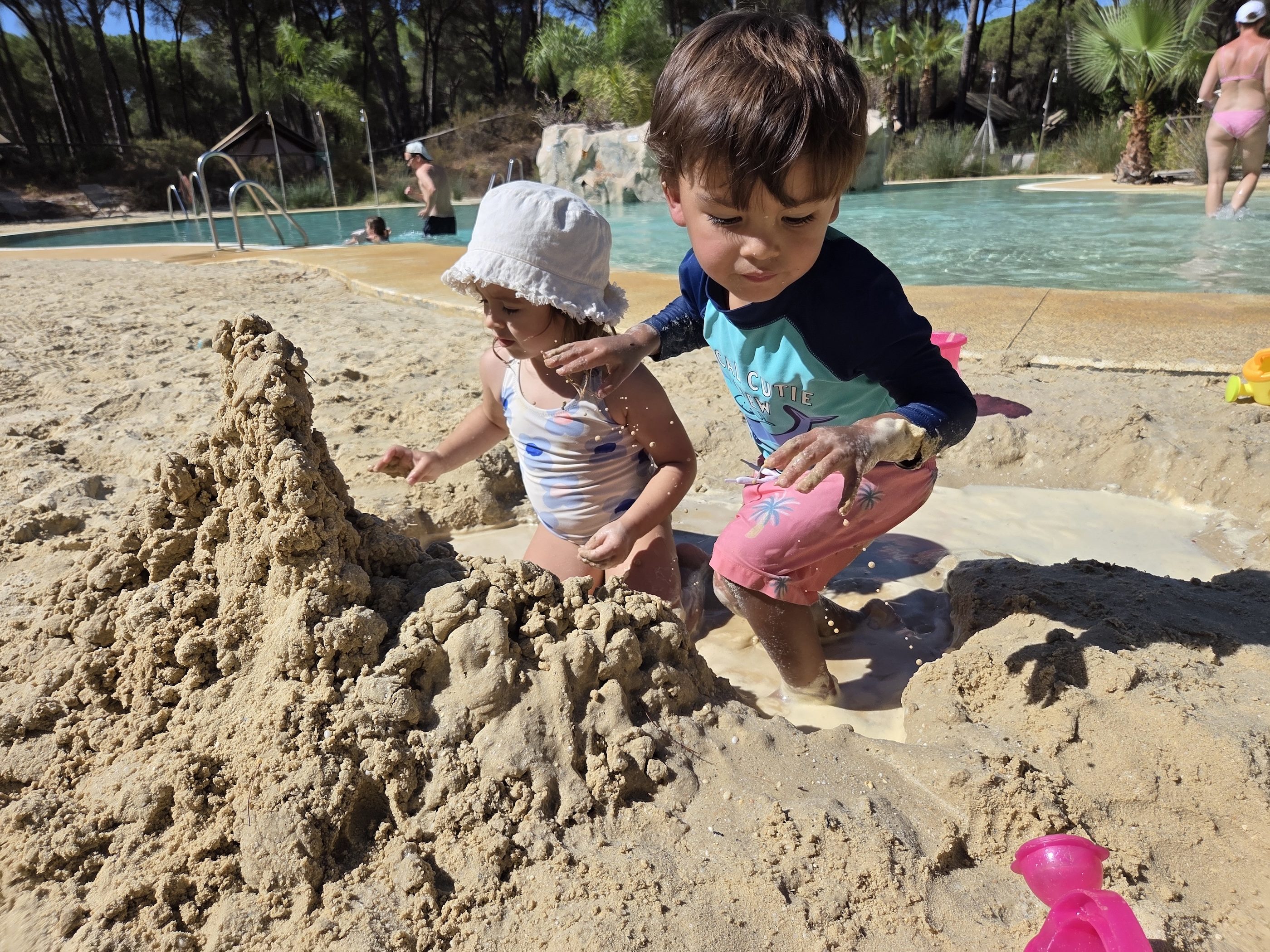 Arlo in a teal rash guard and a little girl in a bucket hat digging together in a big pile of wet sand beside the pool