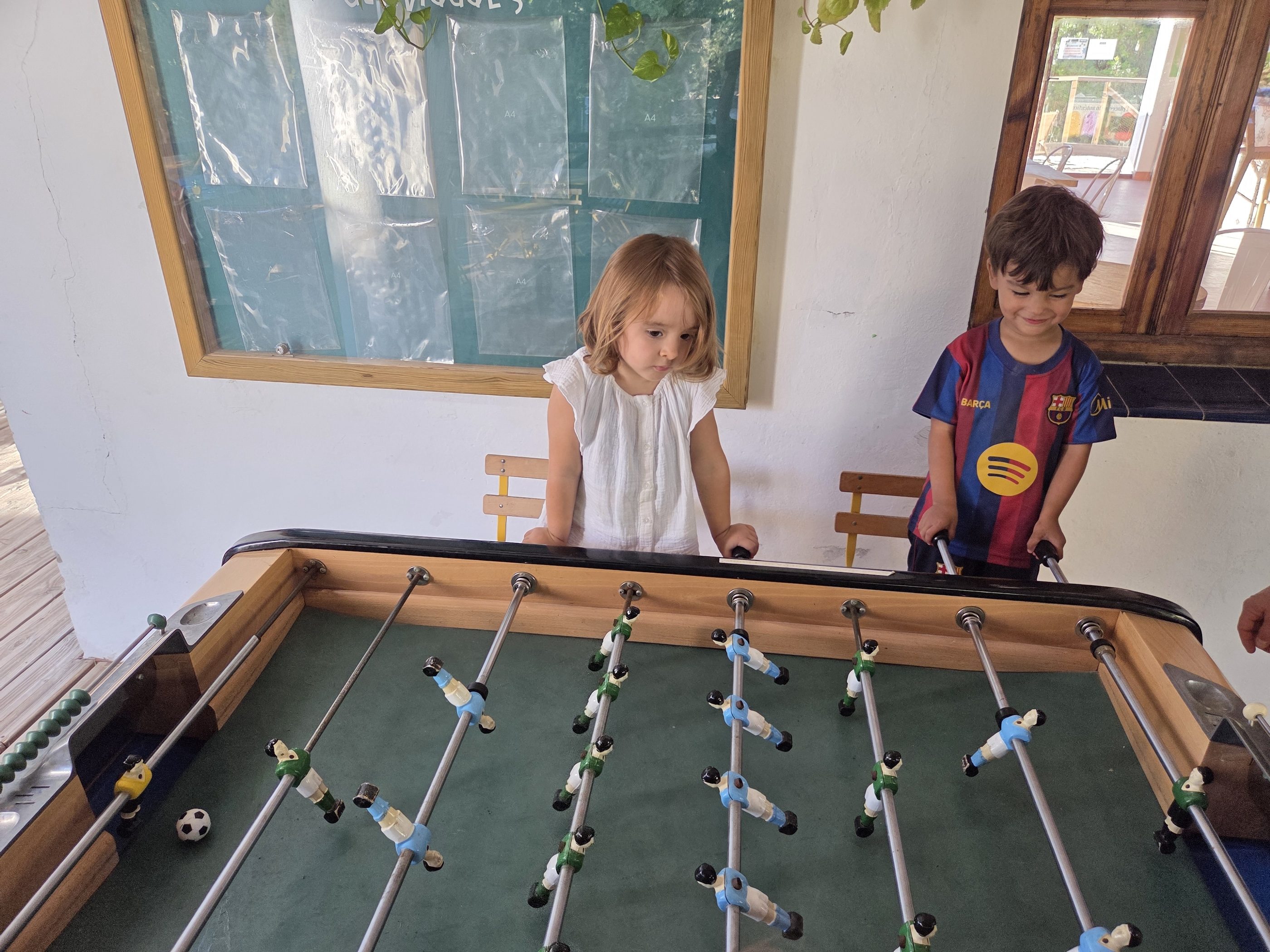 Arlo in his Barça jersey and his new friend in a white dress playing at a wooden foosball table in a whitewashed room