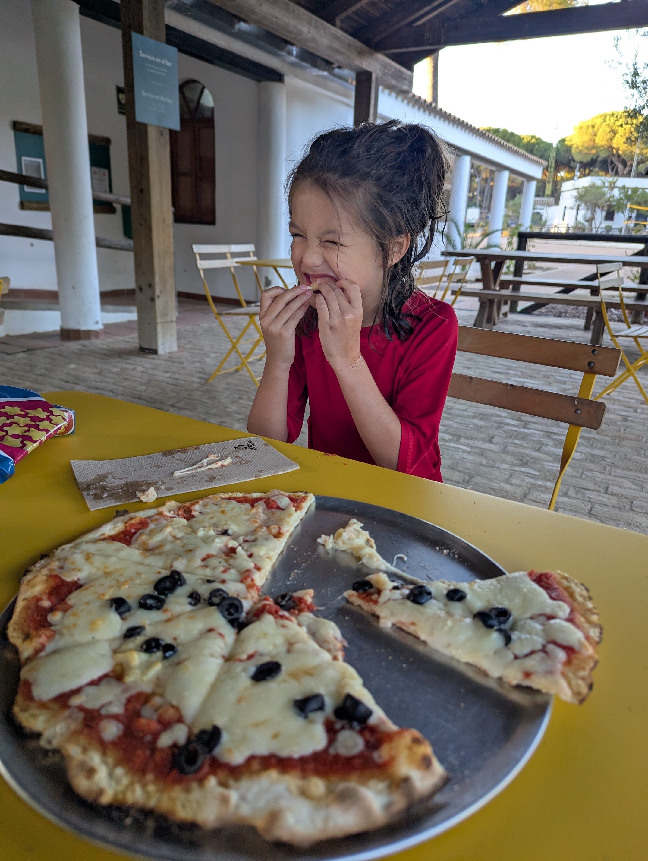 Remi with damp hair in a red shirt grinning with a bite of pizza at a yellow table, a pizza with black olives and melted cheese on a tray in front of her