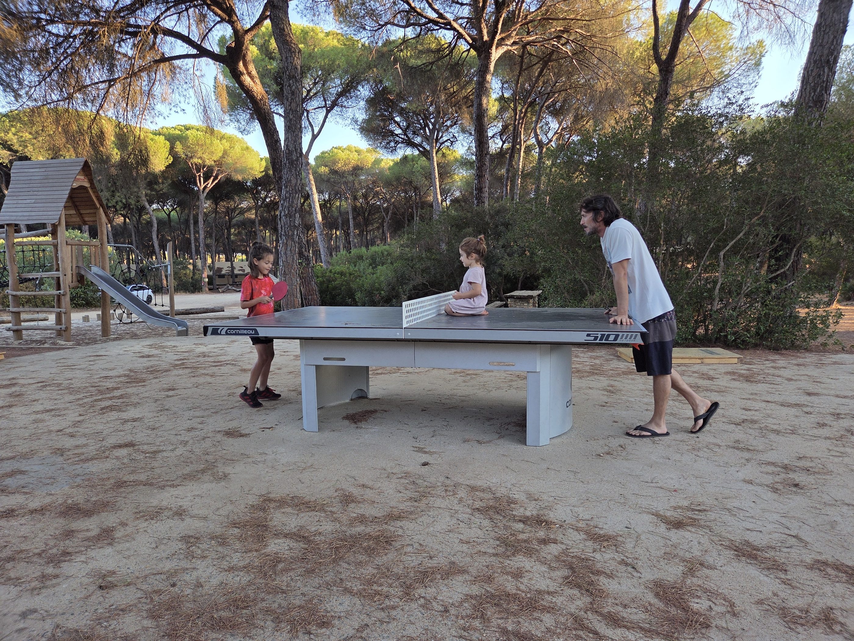 A concrete ping pong table in the sand beside a wooden playground, with Remi on one end, a small child sitting on the table and a parent at the other end
