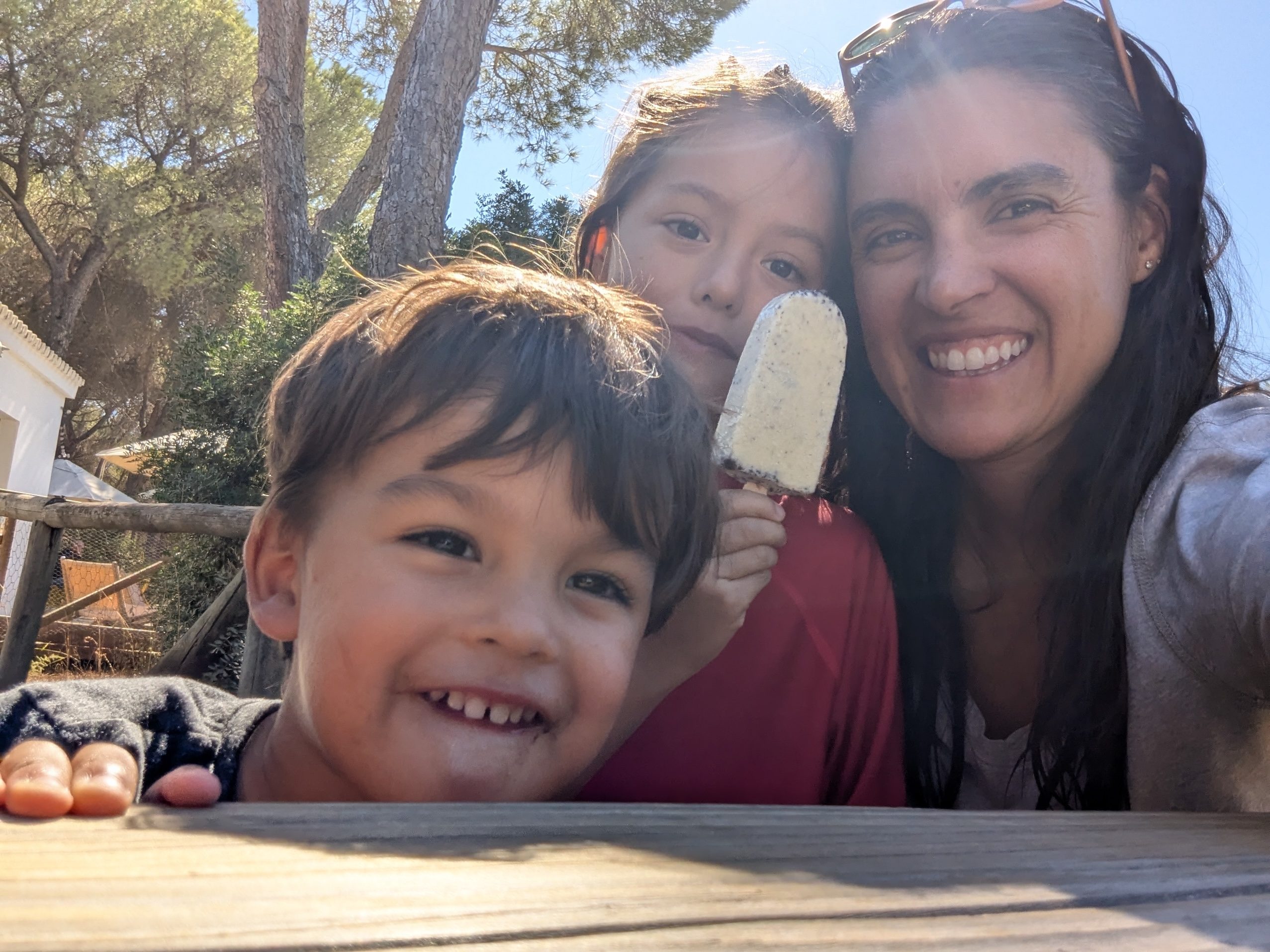 A parent, Remi and Arlo squeezed into a selfie, Remi holding a vanilla popsicle, Arlo grinning at the camera, pine trees behind