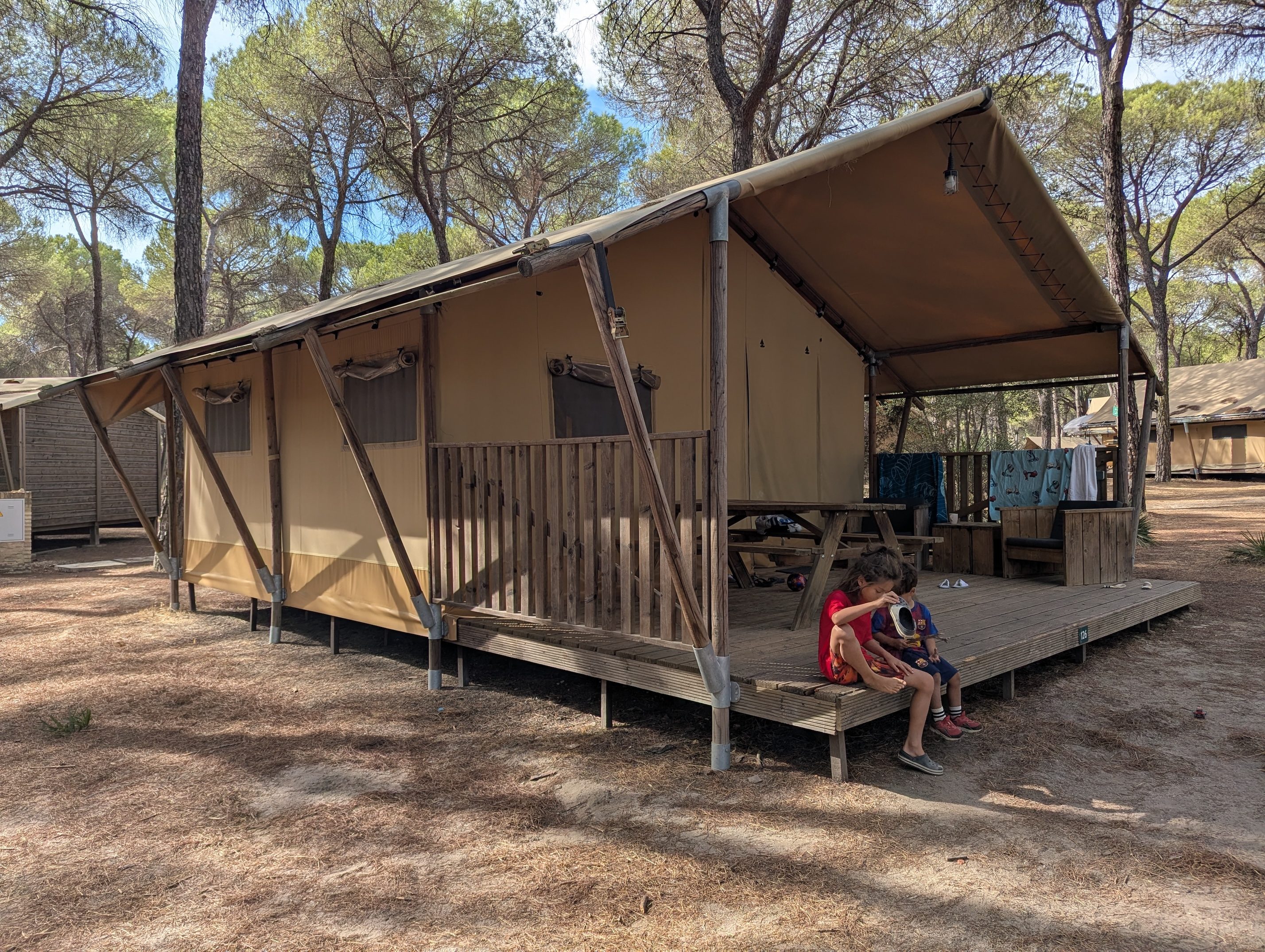 The side of the canvas safari tent on its wooden deck among tall pine trees, with Remi and Arlo sitting on the front steps