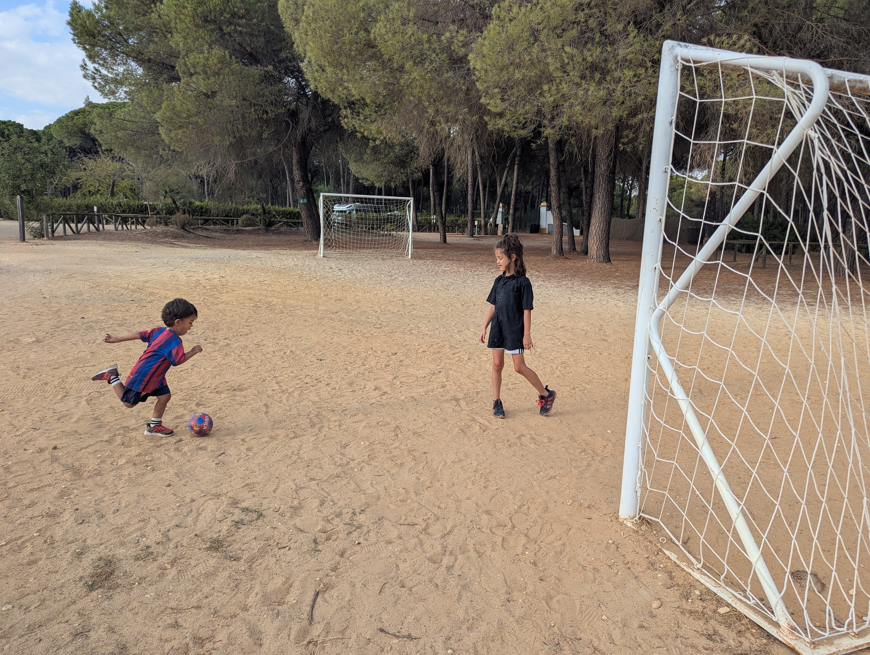 Arlo in his Barça kit kicking a red ball across a sandy soccer field while Remi runs after it, a goal net and pine trees behind them