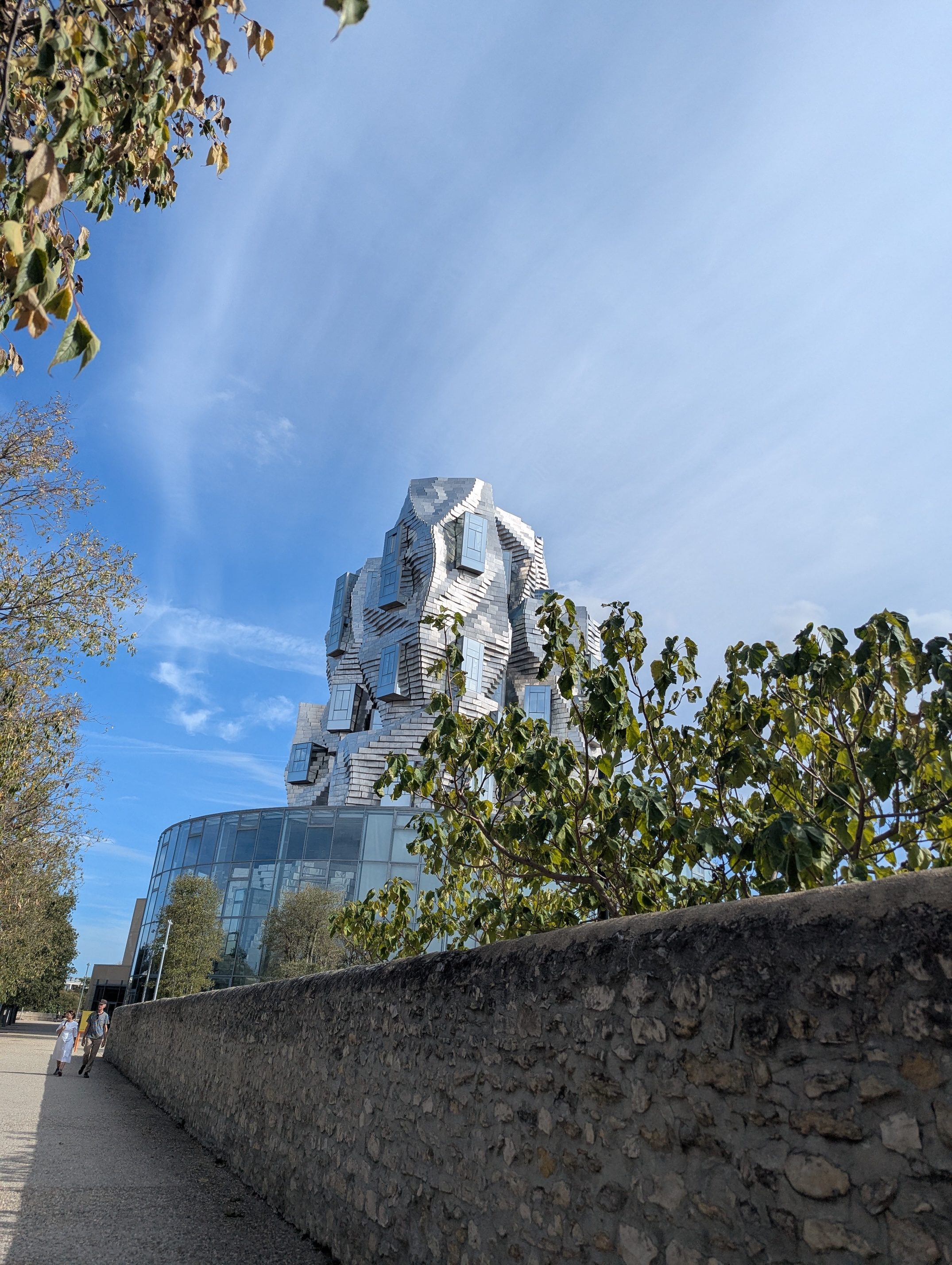 The twisting silver stainless steel tower of LUMA Arles glinting above a stone wall and trees against a blue sky streaked with cloud