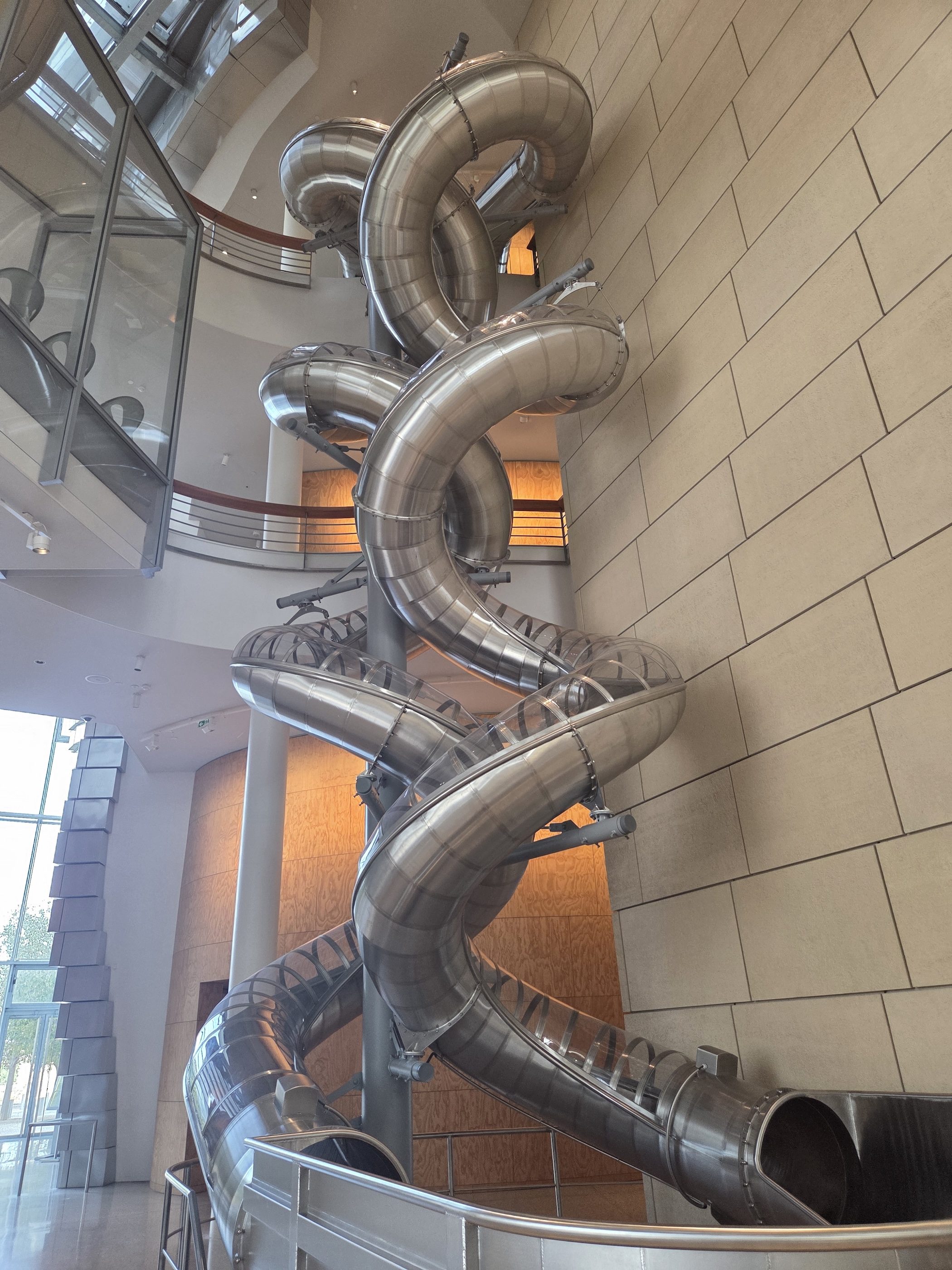 Silver tube slides spiraling down the inside of the LUMA tower atrium past a glass wall and stone-clad panels
