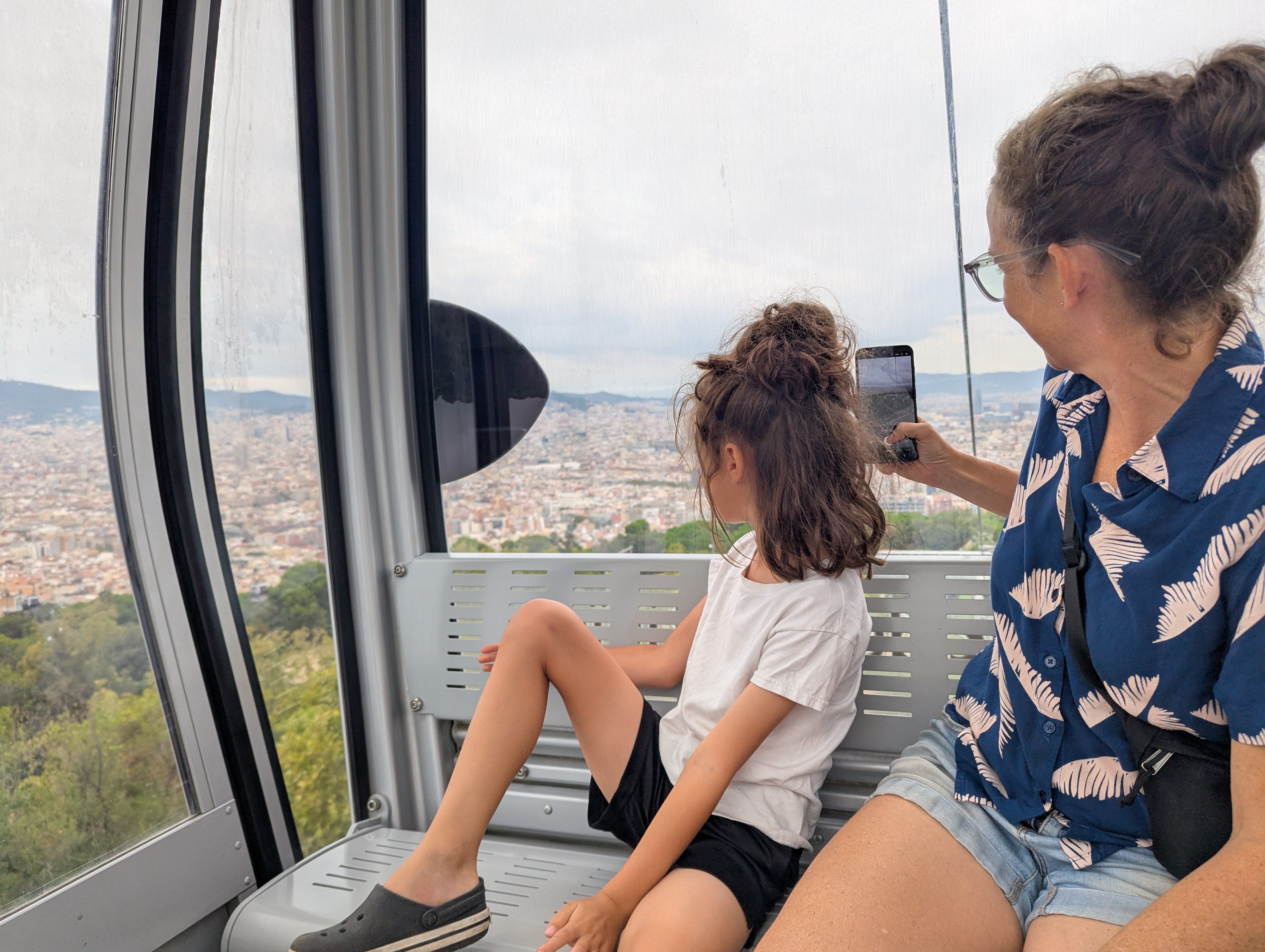 Remi sitting on the bench of a cable car cabin looking out over the sprawl of Barcelona while Dani takes a photo with her phone