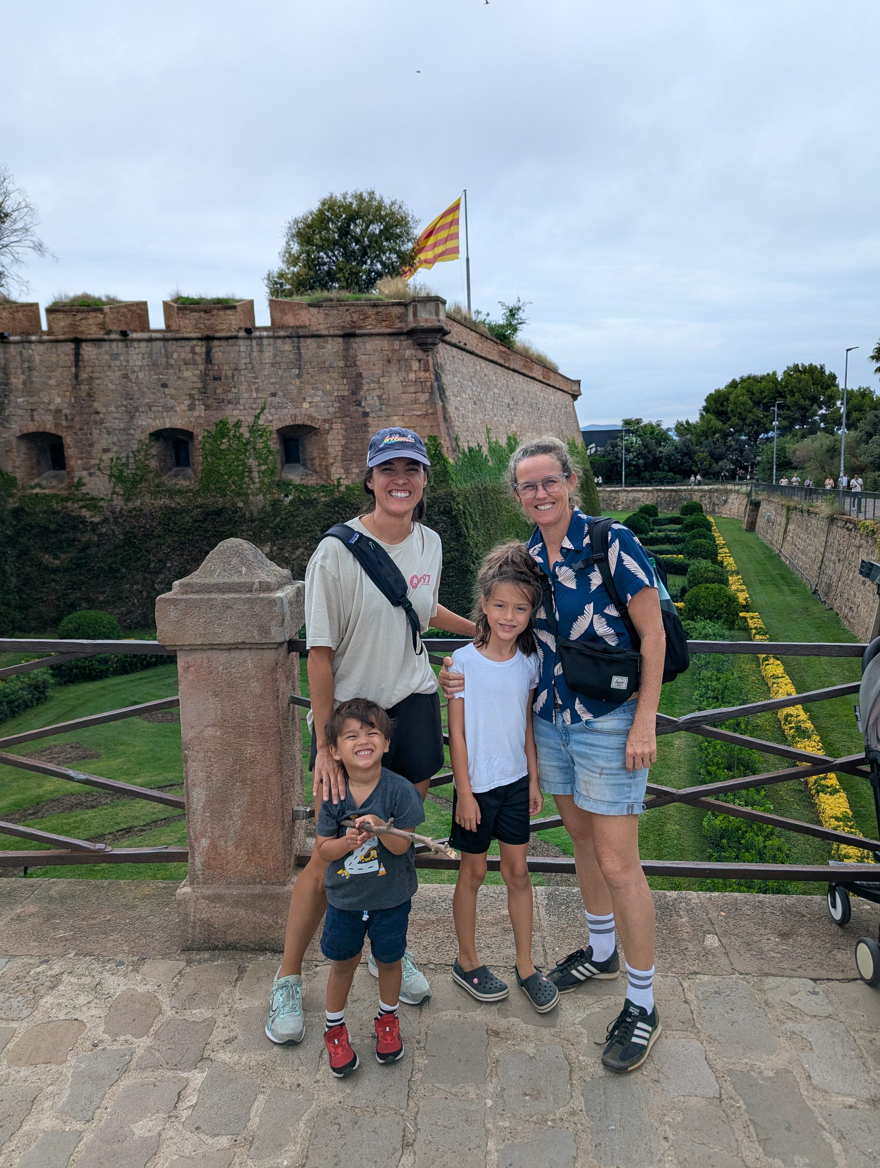 Two adults, Remi and Arlo standing at a railing in front of the stone walls of Montjuïc Castle, a Catalan flag flying behind them