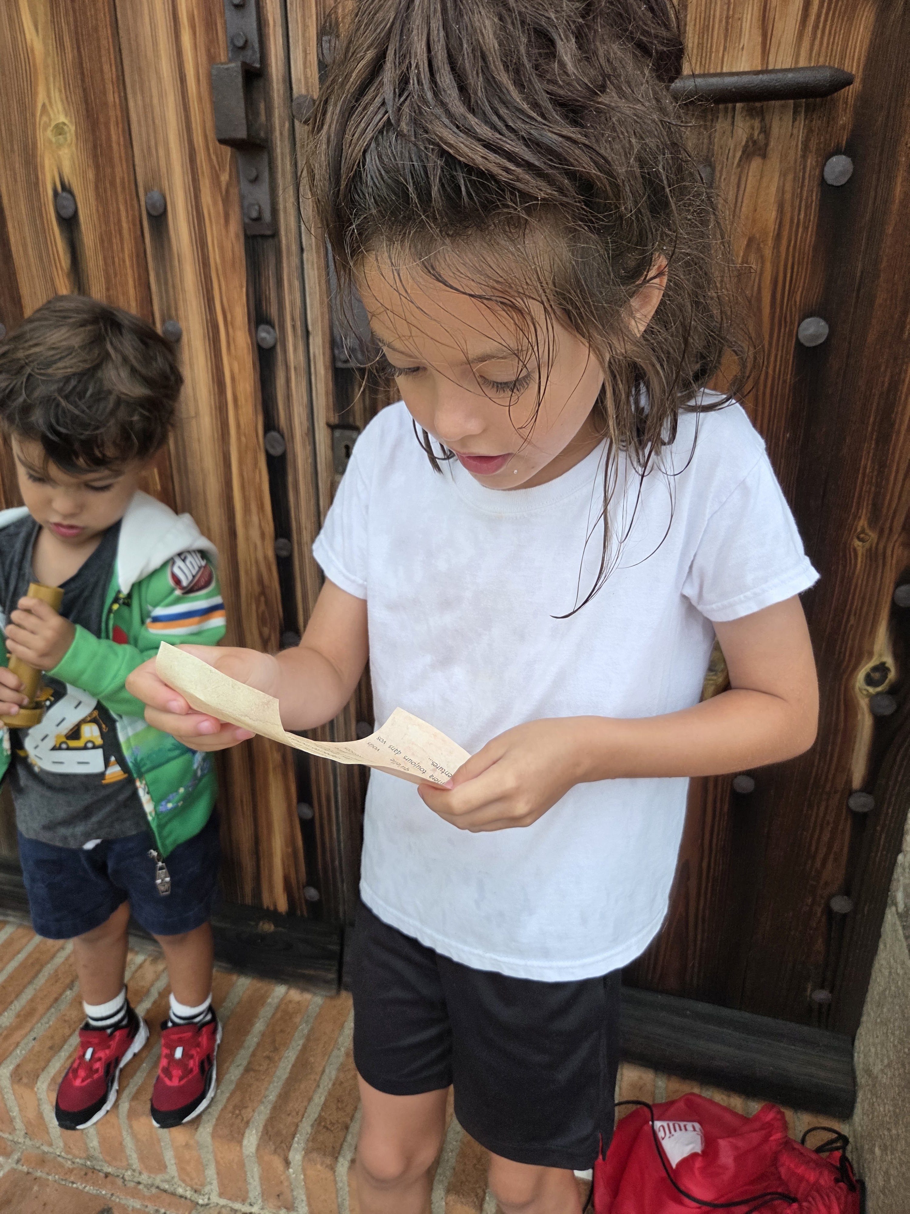 Remi reading a clue on a slip of paper beside Arlo in front of a big wooden door at Montjuïc Castle