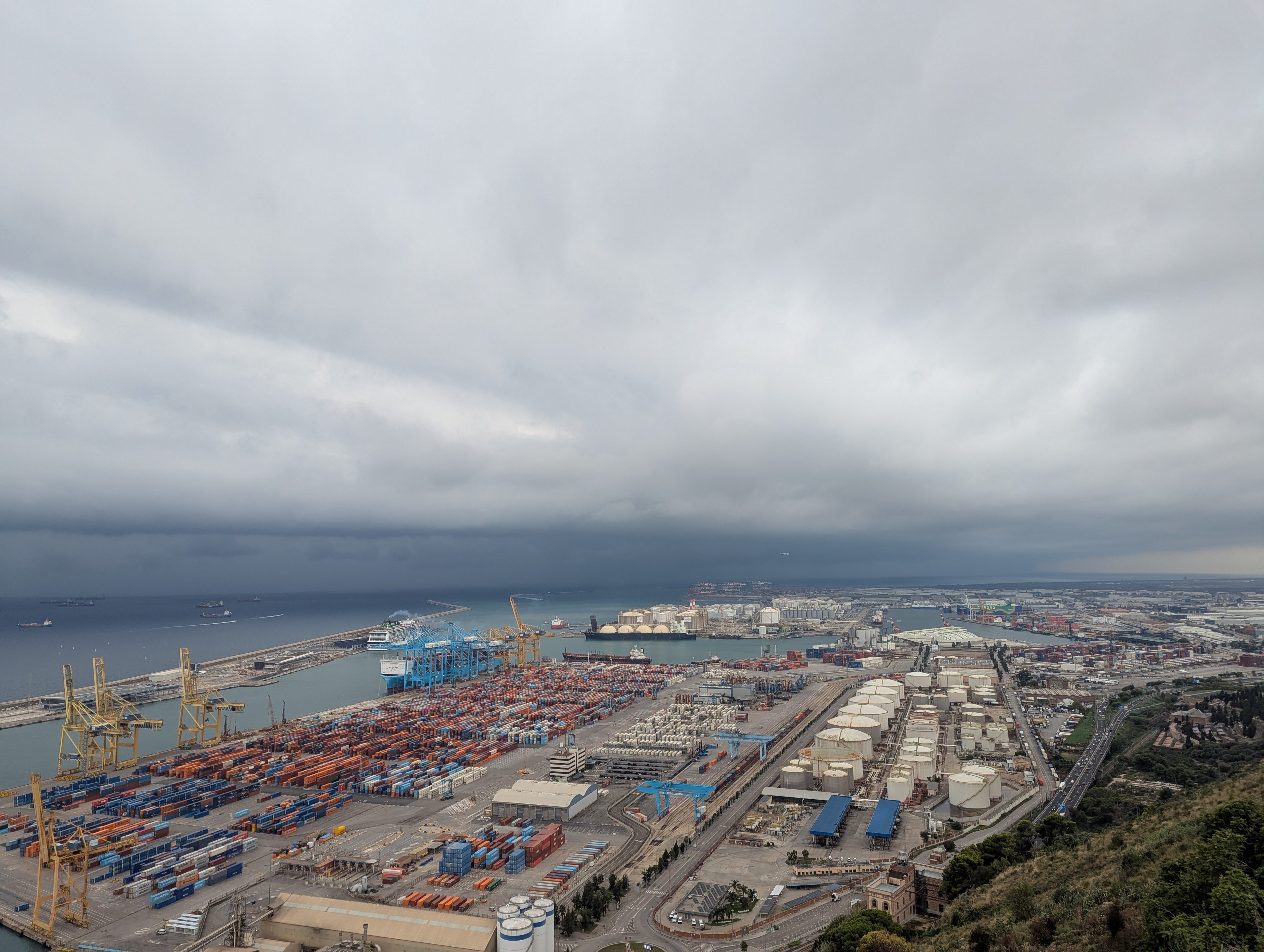 A view from Montjuïc hill over Barcelona's container port, cranes and stacked shipping containers under a heavy gray sky