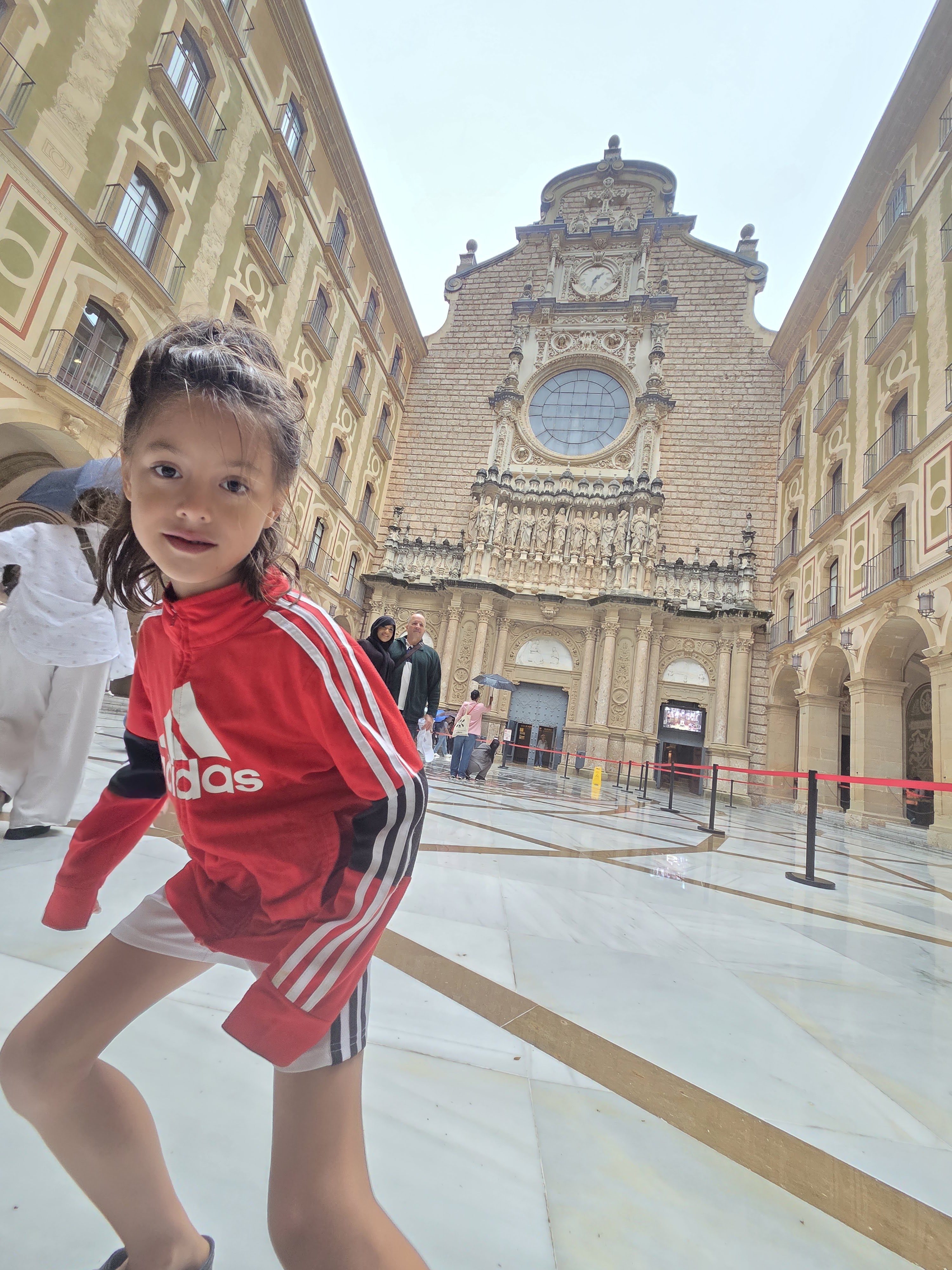 Remi in a red Adidas jacket jumping in front of the Montserrat basilica facade and its rose window, with the stone courtyard wet from rain