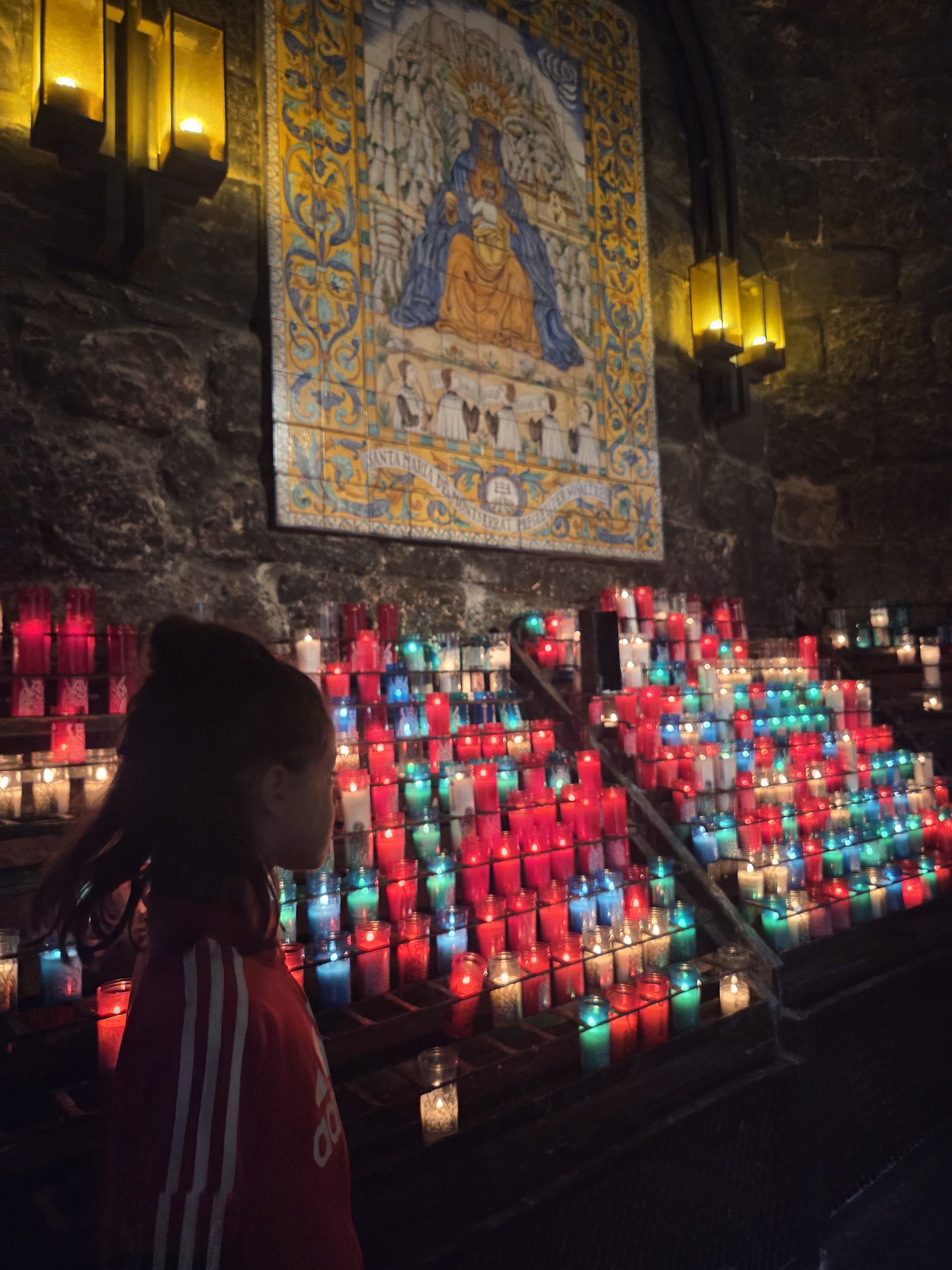 Remi in a red jacket standing in front of rows of red, blue and green candles lit beneath a tiled image of the Virgin in the Montserrat basilica