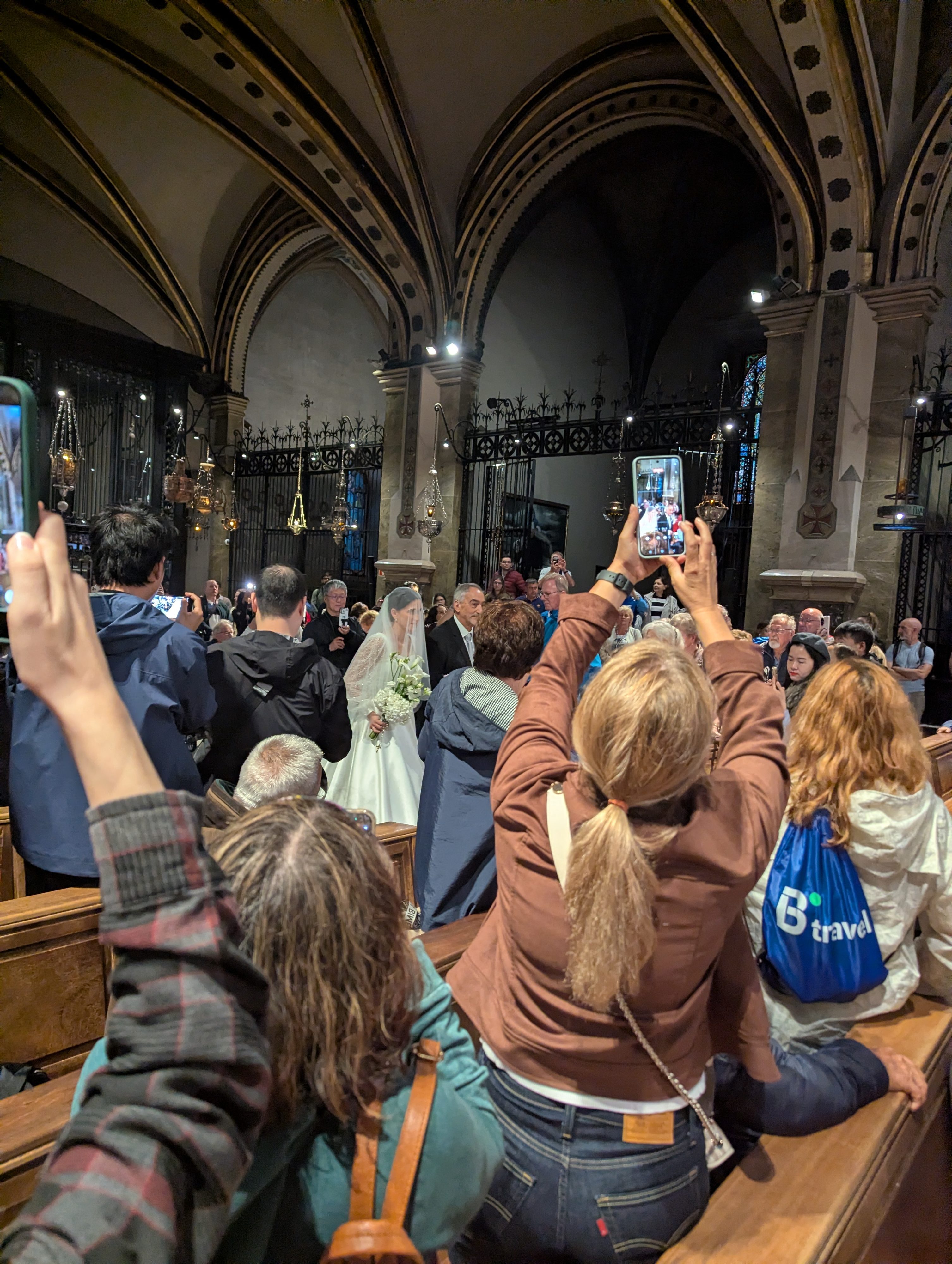 Tourists in raincoats and ponchos holding up phones in the pews of the Montserrat basilica while a bride in white walks down the aisle in the distance