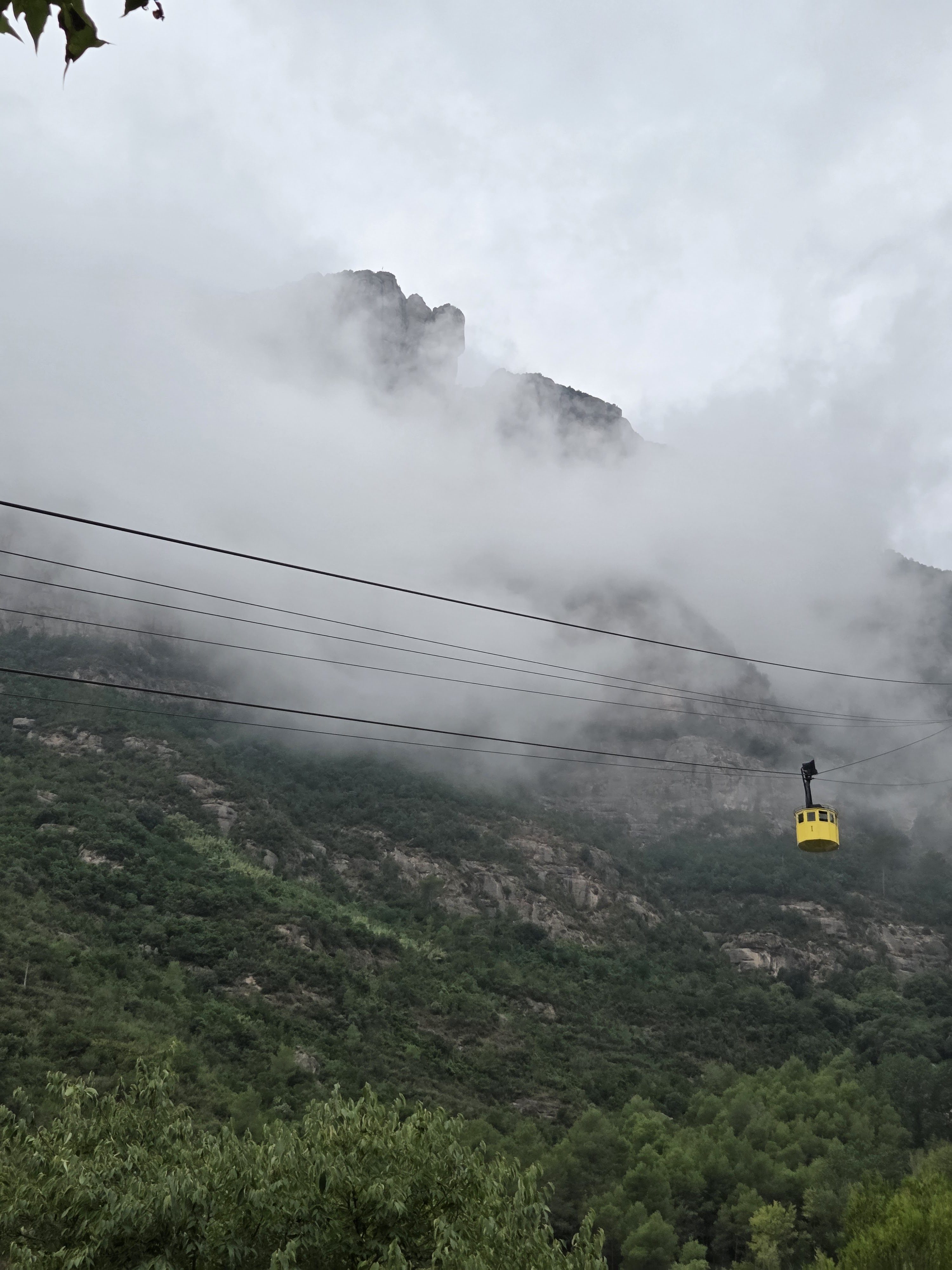 A yellow cable car gondola on its cables above green forested hills, mist hanging over the mountain peaks