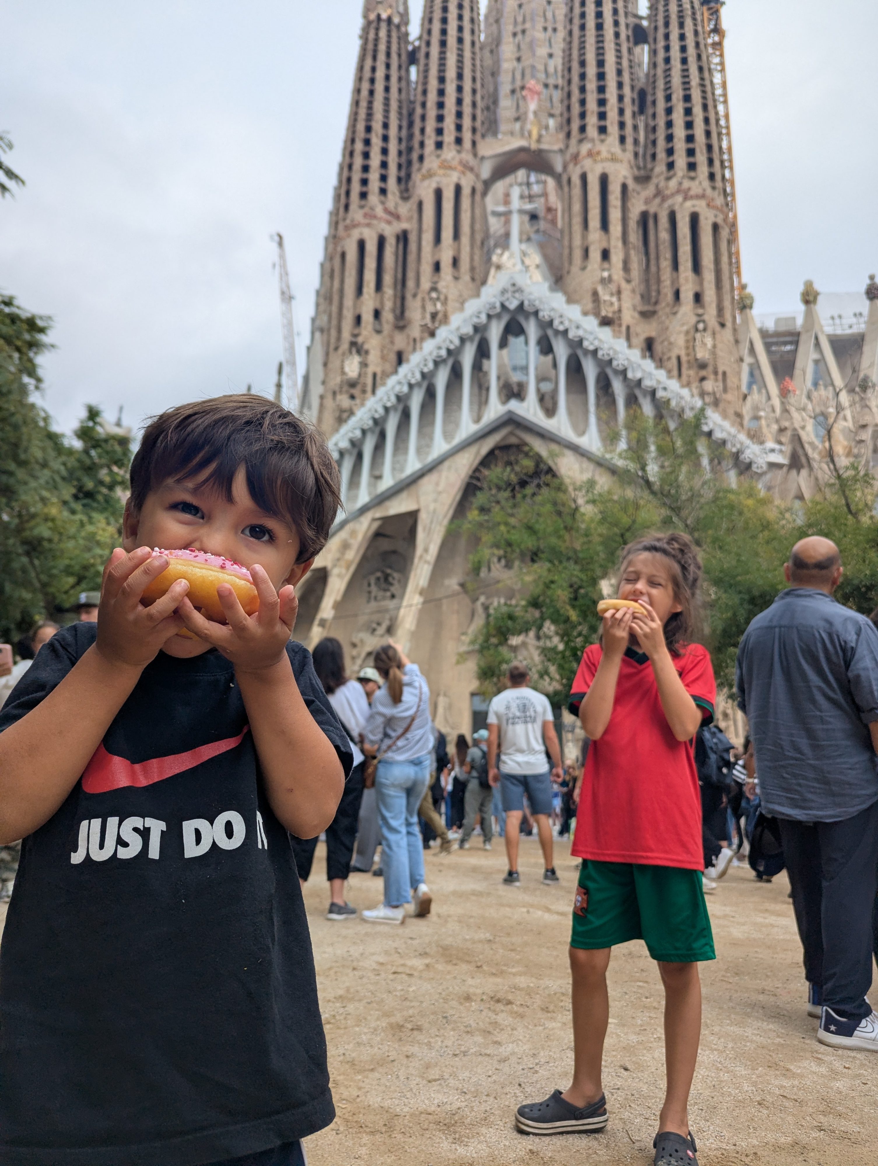 Arlo in a black Nike t-shirt taking a bite of a pink-frosted doughnut while Remi in a red jersey eats one behind him, the Sagrada Família and a crowded plaza in the background