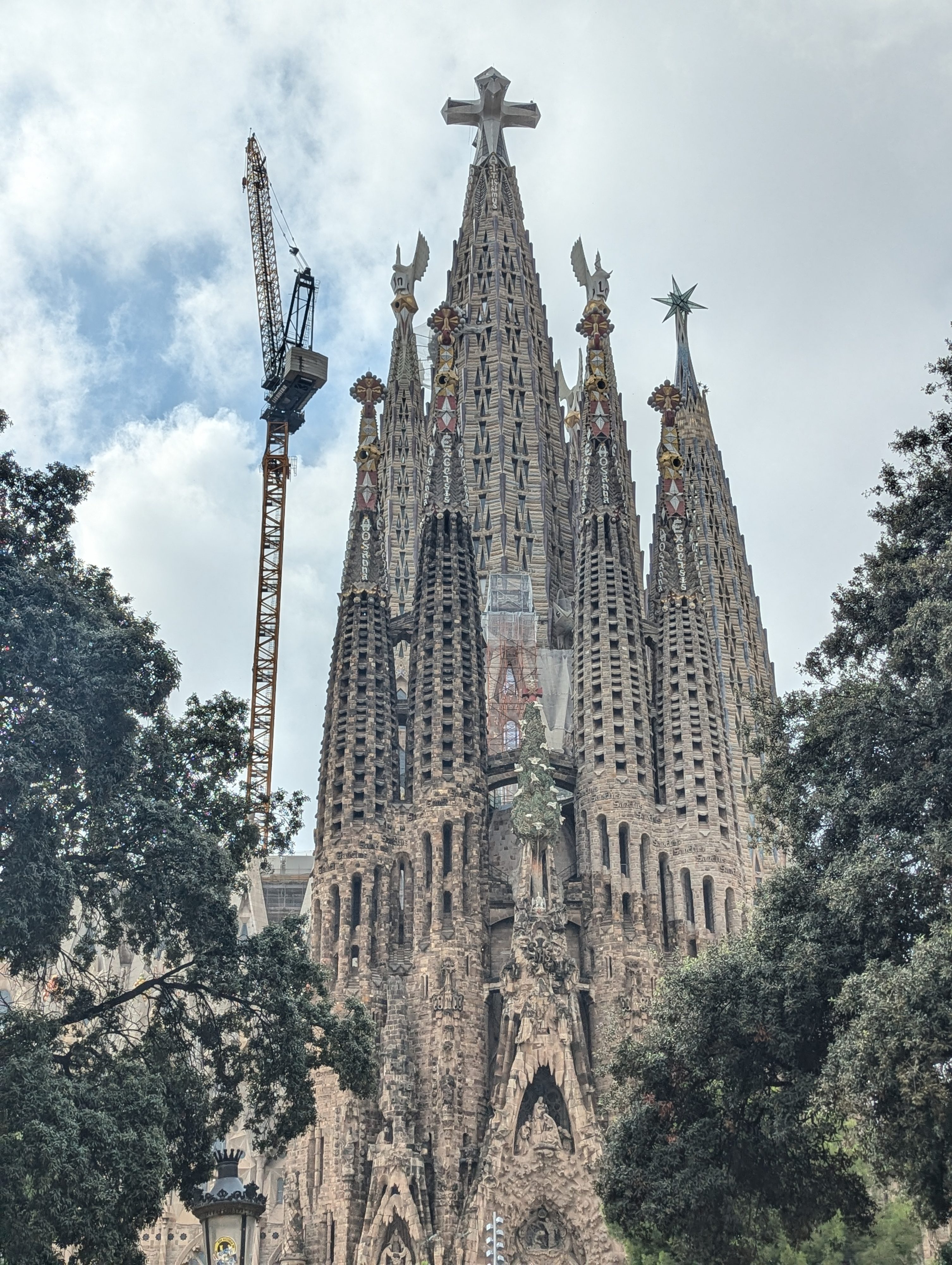 The tall stone spires of the Sagrada Família rising above dark green trees, with a construction crane beside them and a cloudy sky behind