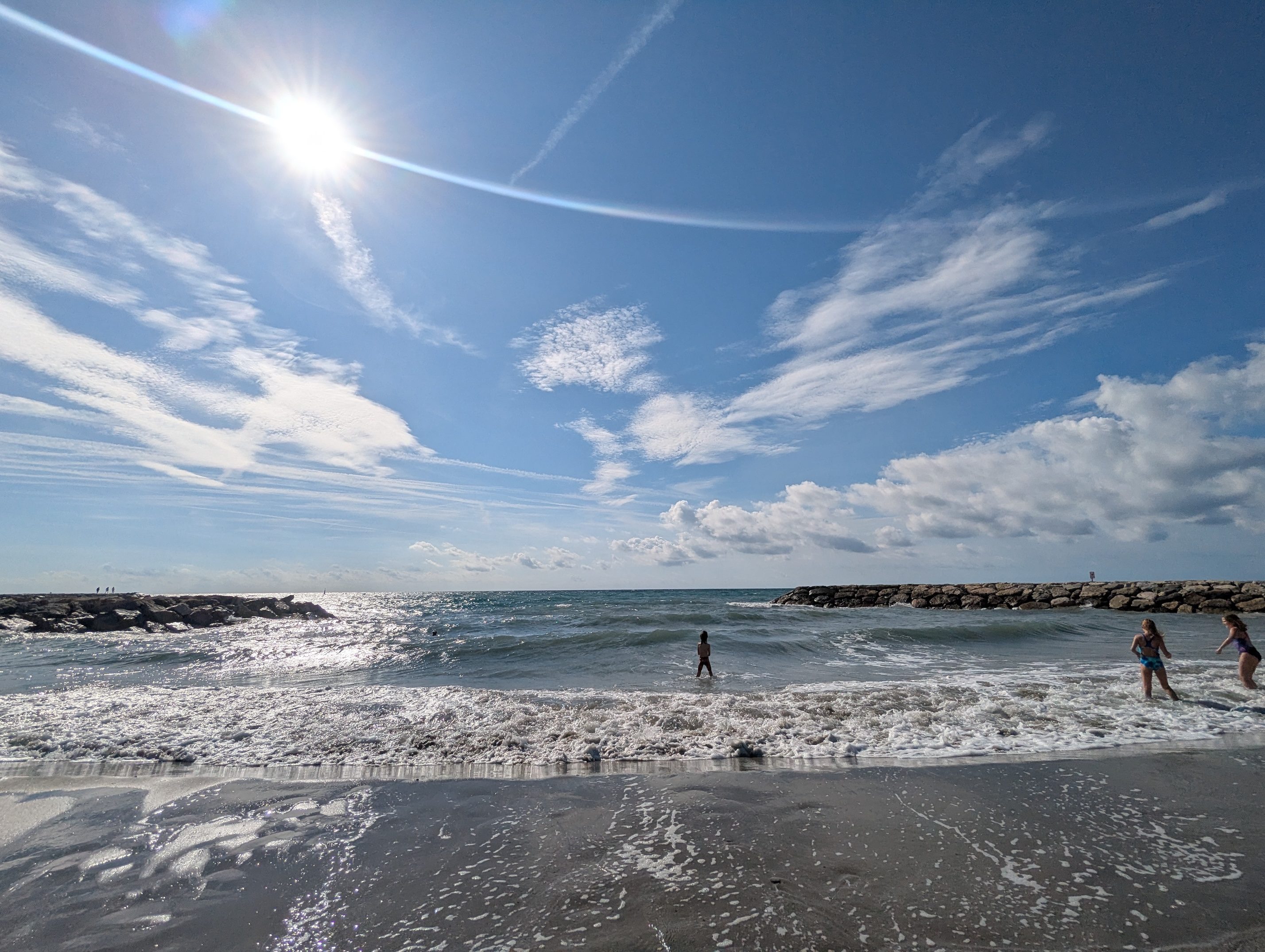 Waves rolling onto the dark sand beach at Saintes-Maries-de-la-Mer, a stone jetty on the horizon and swimmers in the surf under a blue sky streaked with clouds