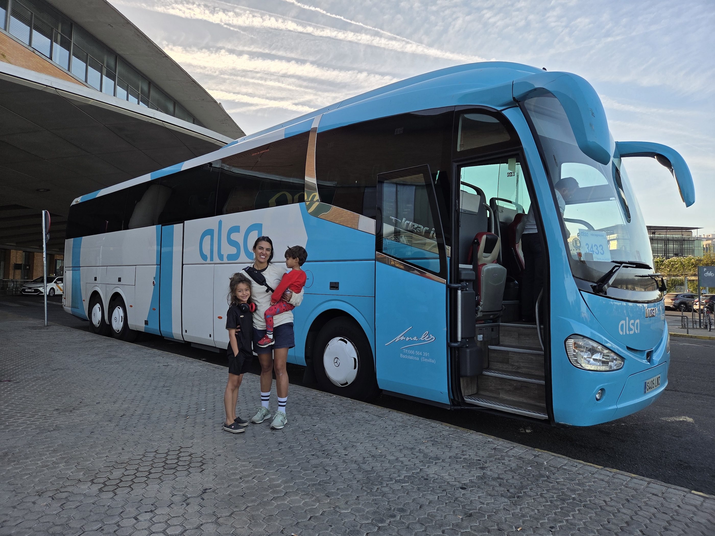 A parent holding Arlo and standing with Remi in front of a light blue Alsa coach at a bus station