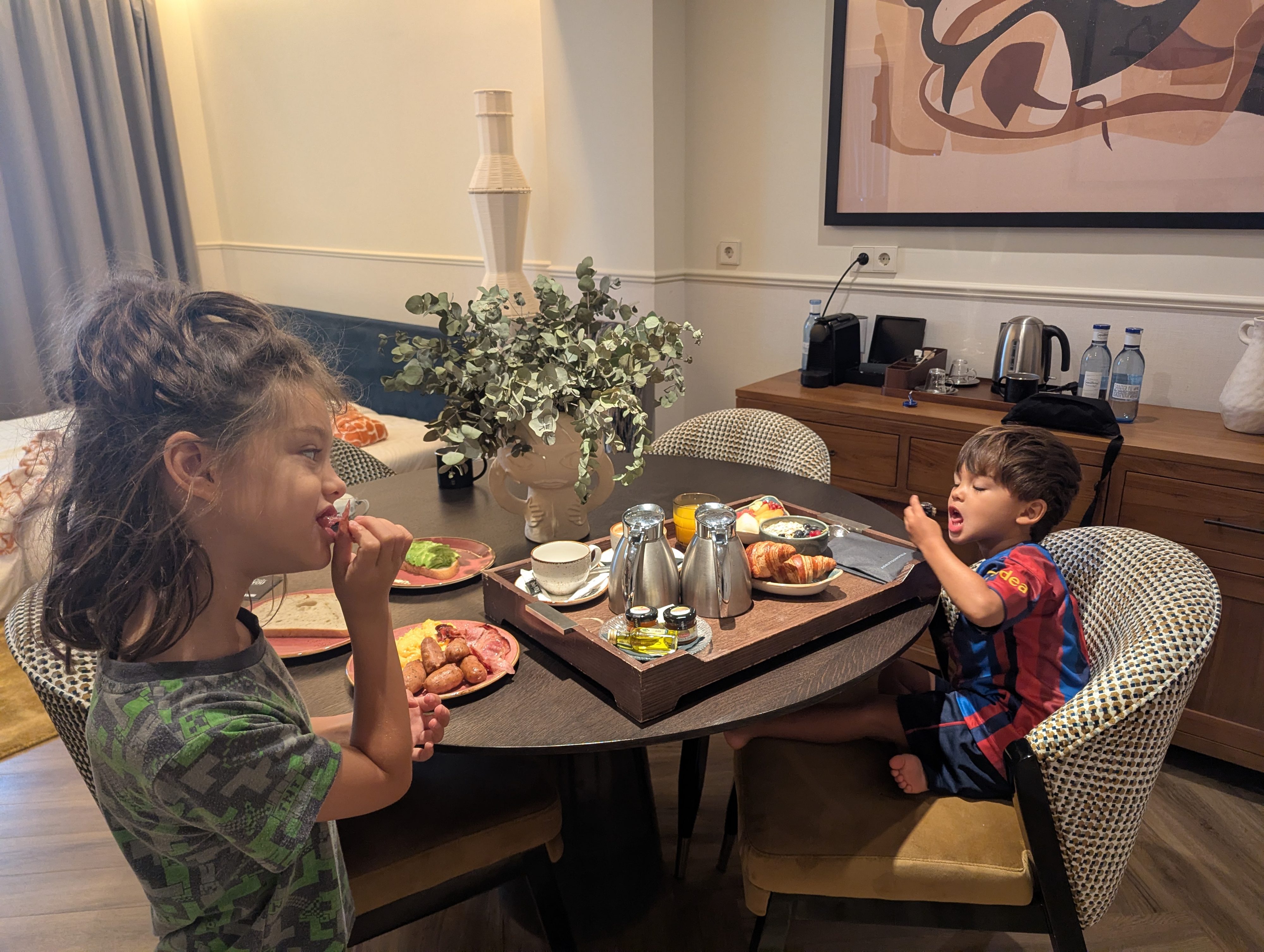 Remi and Arlo eating room service breakfast at a round table in a hotel suite, with a tray of croissants, fruit, eggs and sausages between them